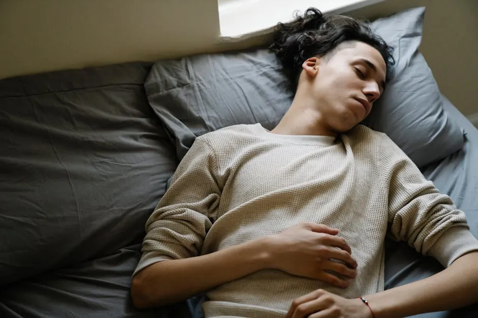 From above of relaxed millennial ethnic male in comfy clothes napping peacefully on soft bed with gray bedclothes