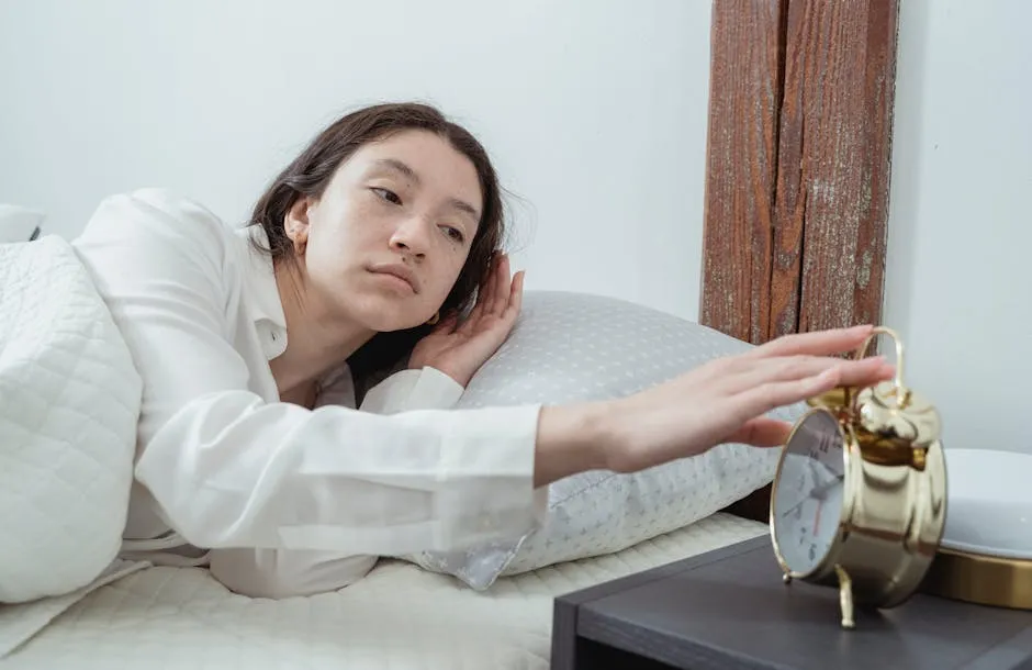 Sleepy young brunette female in white sleepwear resting in comfortable bed and waking up with alarm clock at home in morning time