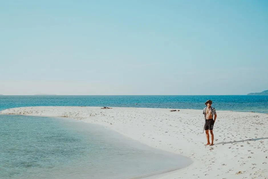 A lone man stands relaxed on a picturesque white sandy beach by clear blue waters, under a bright sky.