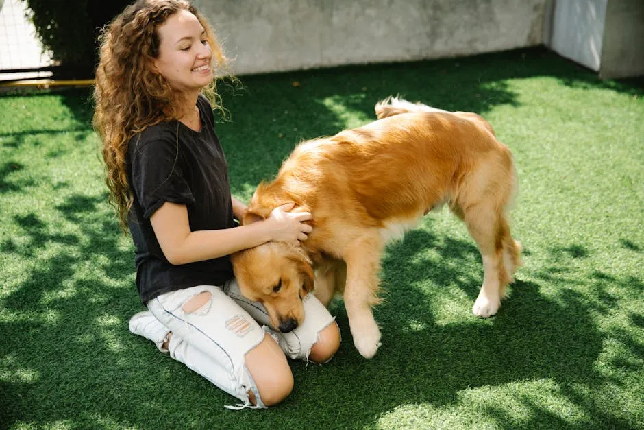 Cheerful young female with curly hair smiling and scratching neck of loyal Golden Retriever dog while kneeling on grass on sunny day in yard