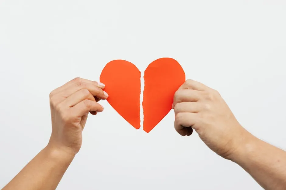 A close-up view of hands holding a torn red paper heart, symbolizing heartbreak or relationship issues.