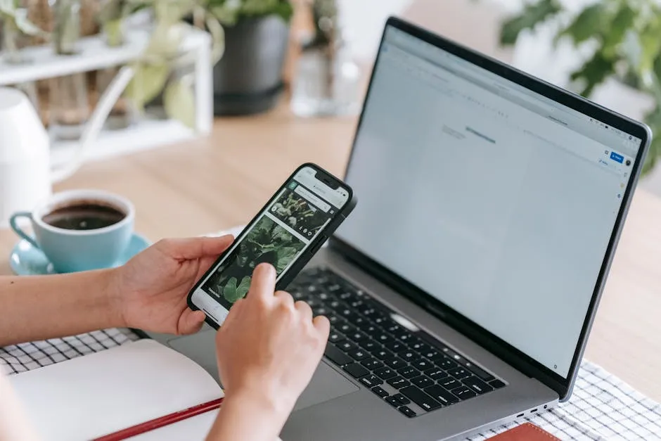 Person using a smartphone and laptop at a desk with coffee and plants, epitomizing modern remote work.