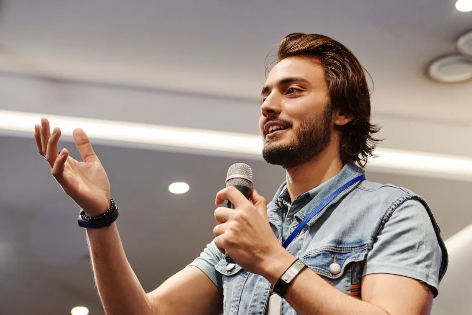 Young man with microphone speaking confidently at an indoor conference, gesturing with a smile.