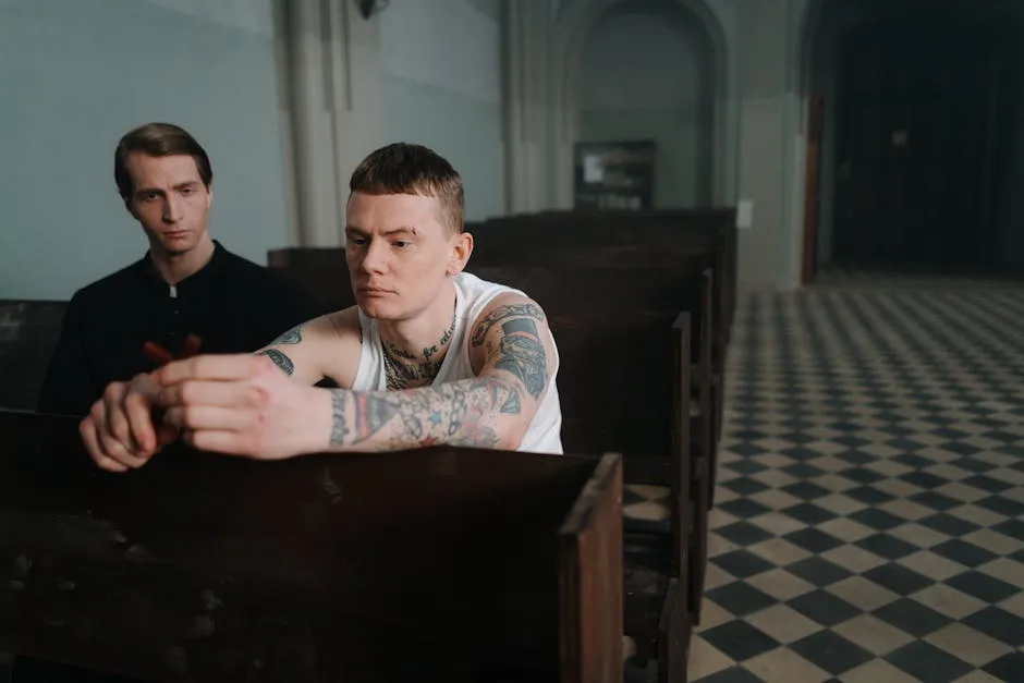 A tattooed man sits pensively in a church pew, holding a crucifix, with a priest nearby.