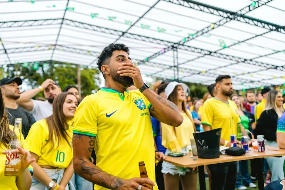 A diverse group of fans in yellow jerseys watch a football match with emotion under a glass roof.